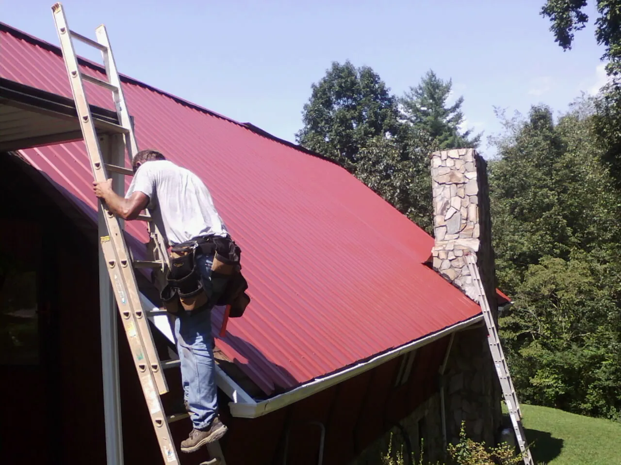 Roofer on a ladder beside a red metal roof and stone chimney — Soffit & Fascia Repair by Roofing Brothers in Burnham